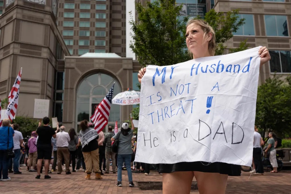 A woman stands at a protest holding a sign reading 