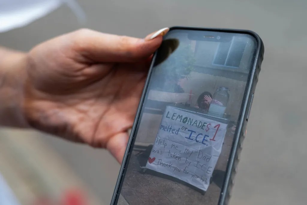 A hand holds a phone displaying a photo of a child selling lemonade with a sign reading, 