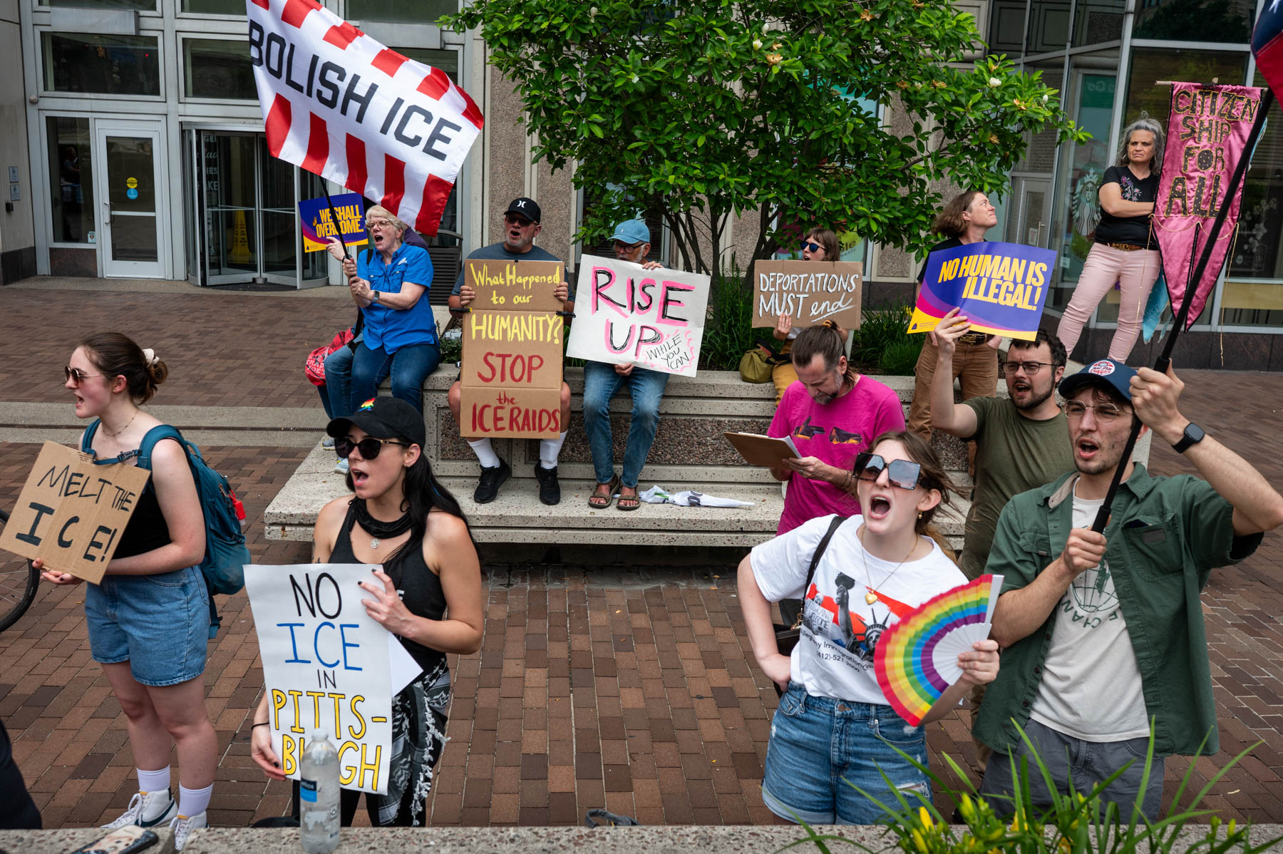 A group of people protest outdoors with signs opposing ICE and deportations; some signs read 