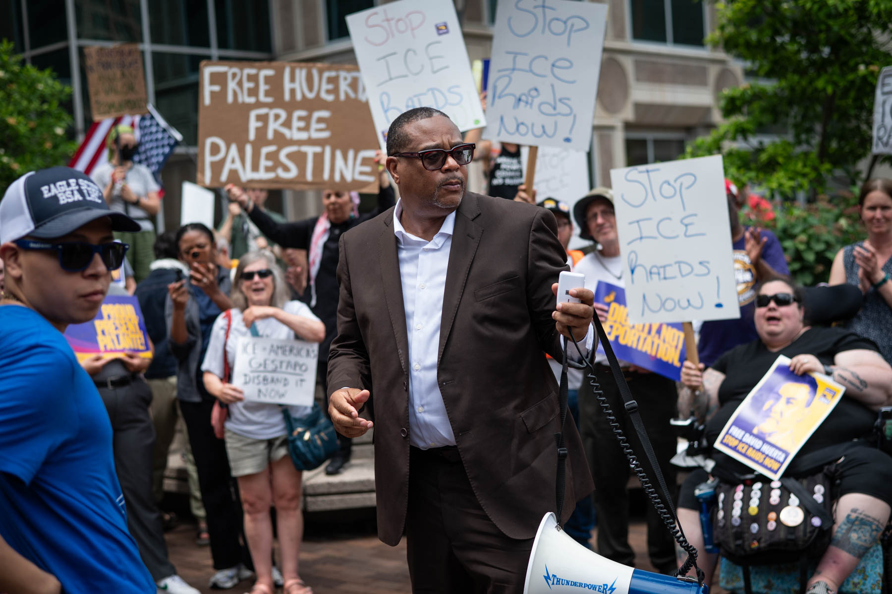 Mayor Ed Gainey in a suit holds a megaphone at a protest, surrounded by people holding signs with messages such as “Stop ICE Raids Now” and “Free Huerta, Free Palestine.”.