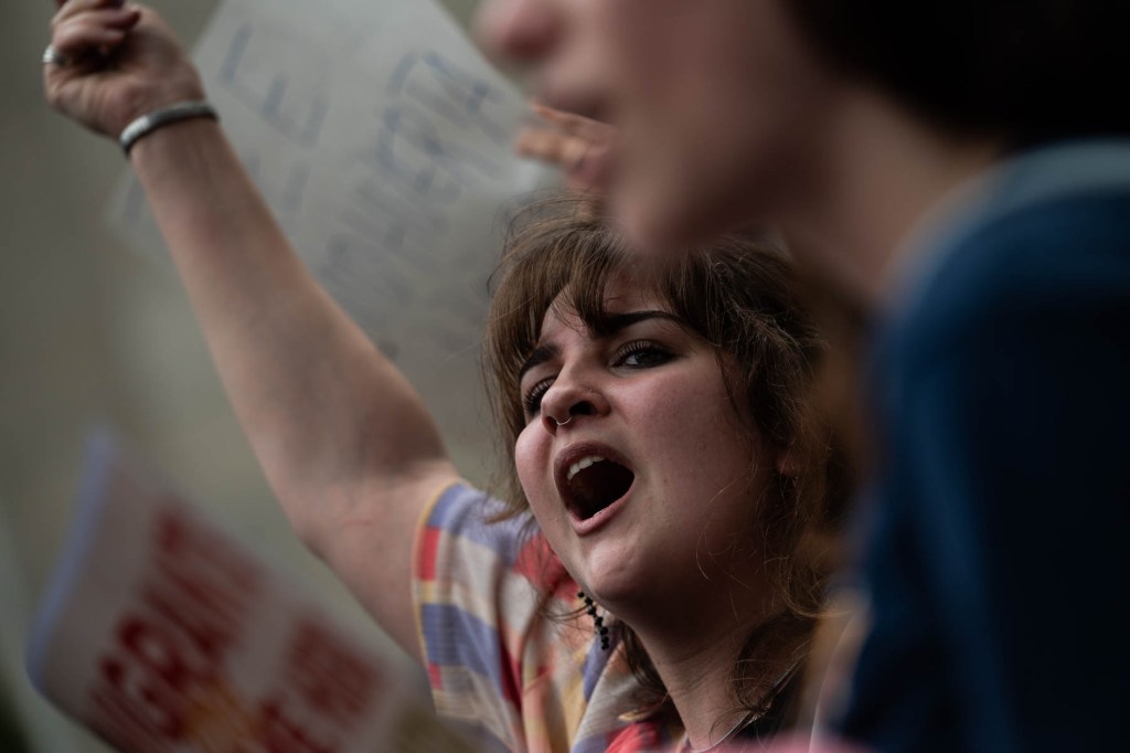 A person with their mouth open raises a fist in the air while holding a sign at a protest. Other people are partially visible in the foreground and background.