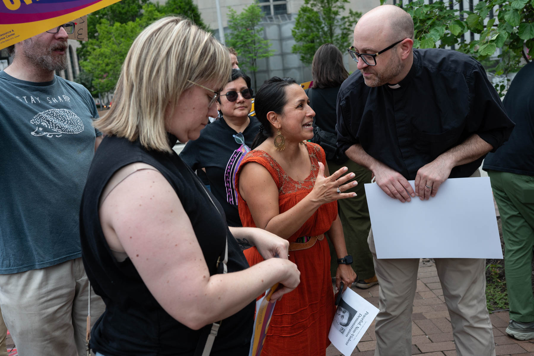 A group of people stand outdoors, engaged in conversation. One woman speaks to a man in clerical attire, while others hold papers and listen. Trees and buildings are visible in the background.